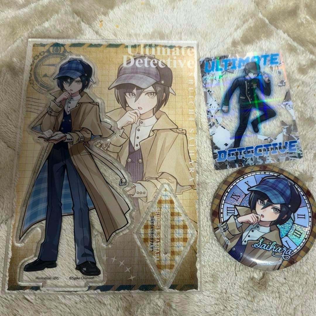 A large acrylic stand of Saihara Shuichi displayed with a matching clear card and a round can badge on a soft surface.