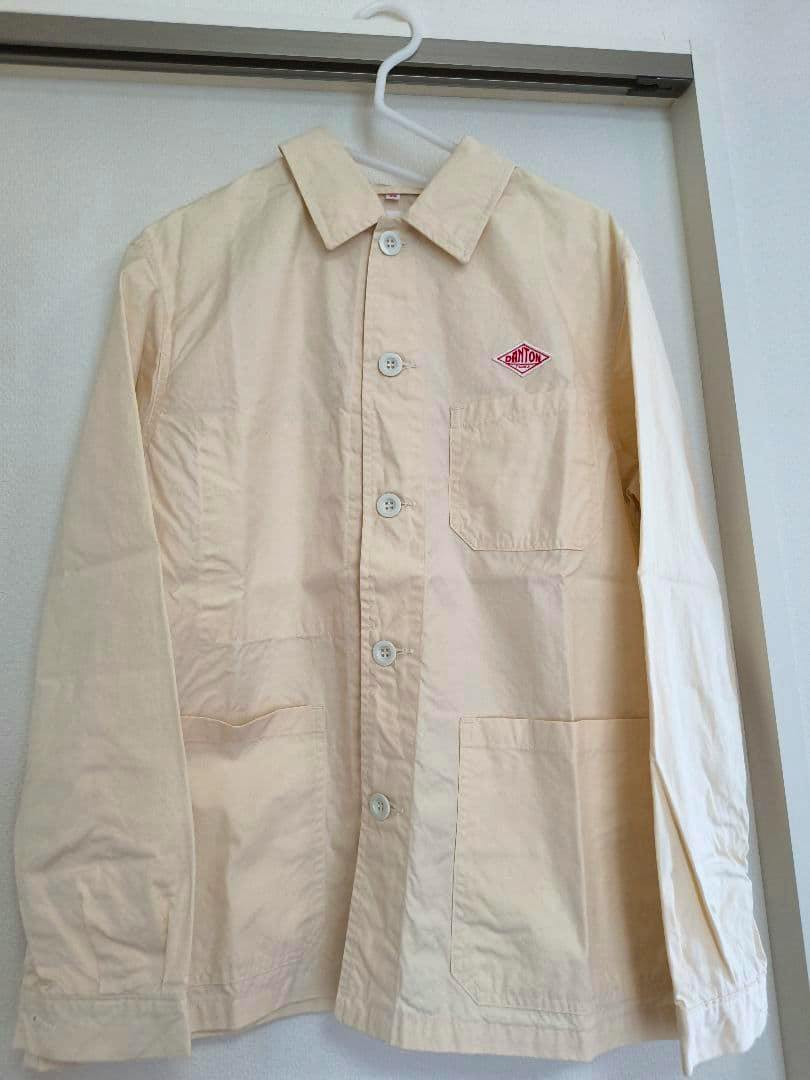 Front view of an ivory DANTON coverall jacket hanging on a hanger, showing buttons, a chest pocket and the DANTON logo.