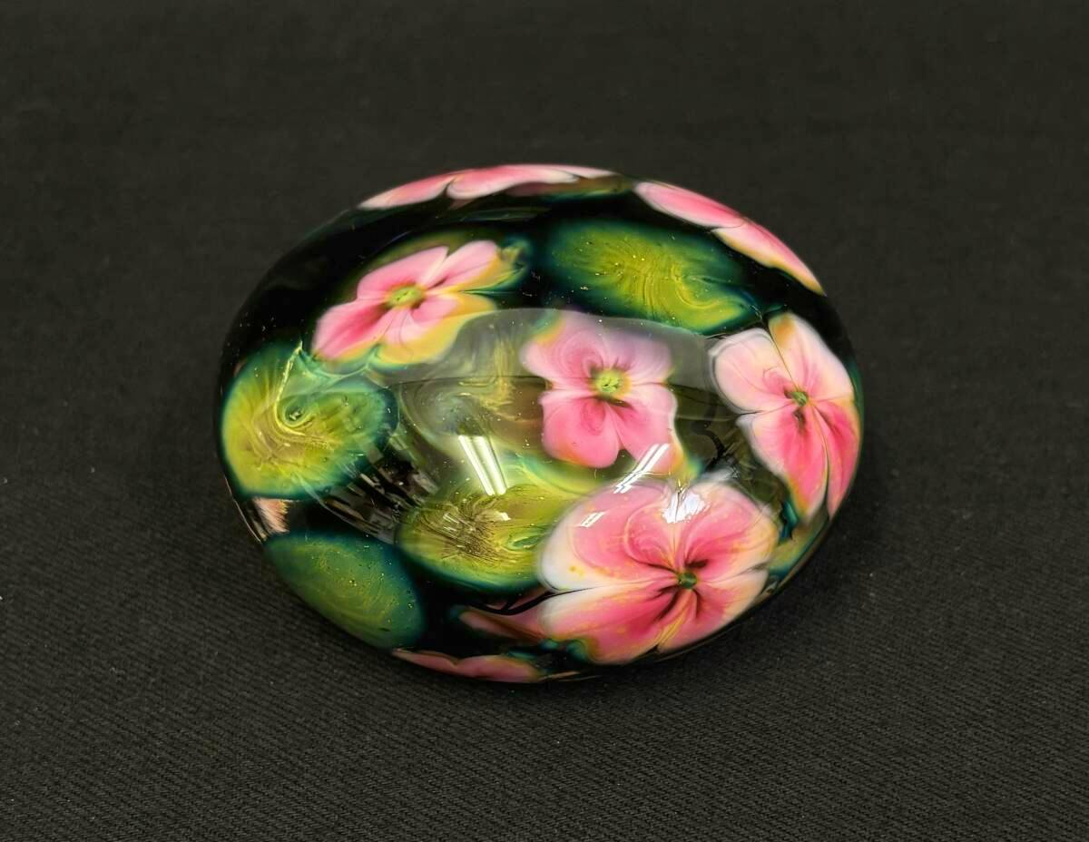A glass paperweight with pink floral motifs and green accents displayed on a dark background.