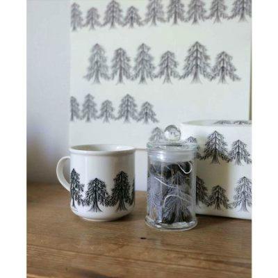 A white ceramic mug with a black forest tree pattern sits beside a glass jar of teabags and matching patterned packaging.