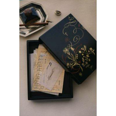 Black lacquered wooden box with gold floral decoration and its lid partially removed, revealing stacked letters inside on a tabletop.