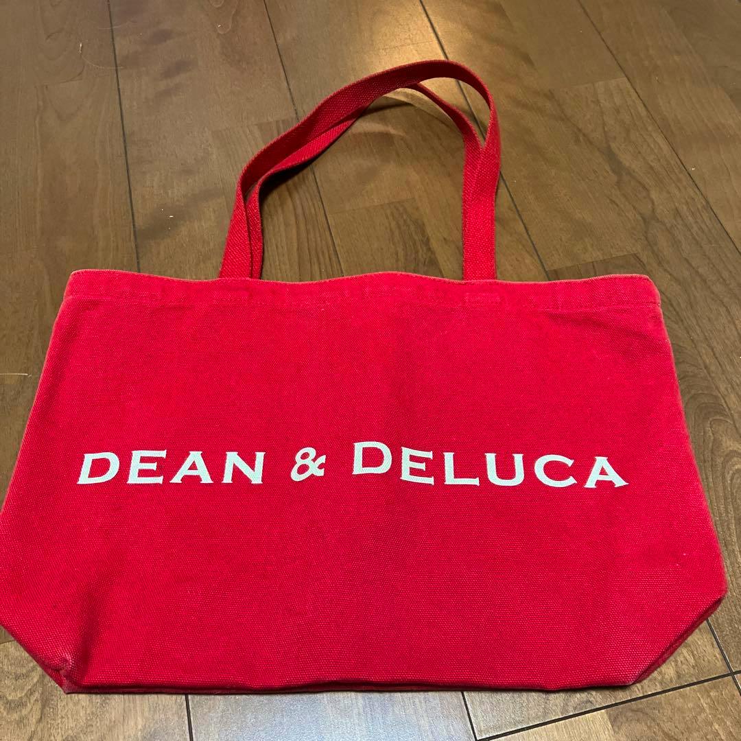 Red DEAN & DELUCA tote bag with shoulder handles lying on a wooden floor.