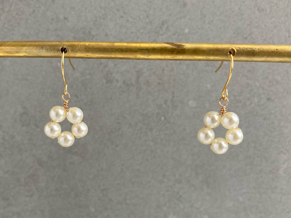 Two dangling flower-shaped pearl earrings with metal hooks hanging from a horizontal rod against a gray background.