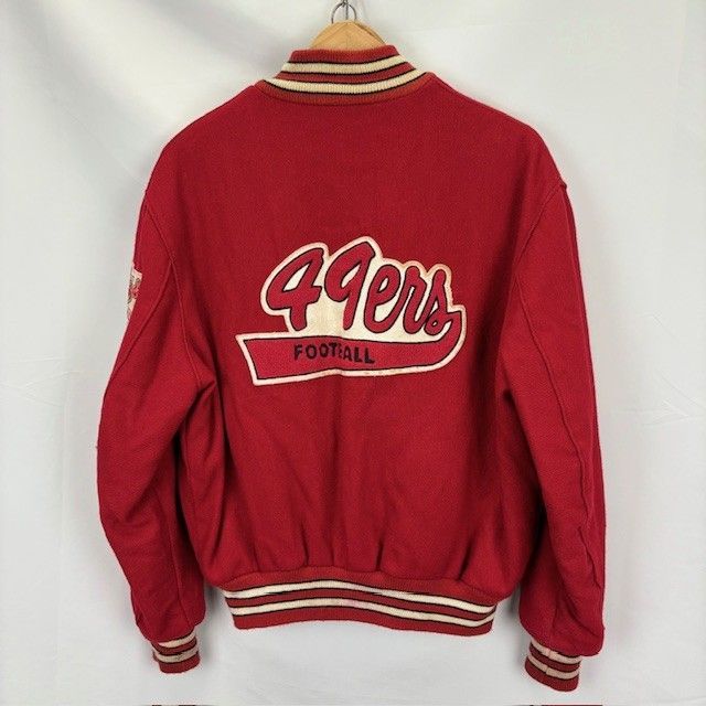 Back view of a men's red wool 49ers jacket with a "49ers Football" back patch, striped collar and cuffs, hanging on a hanger against a white background.