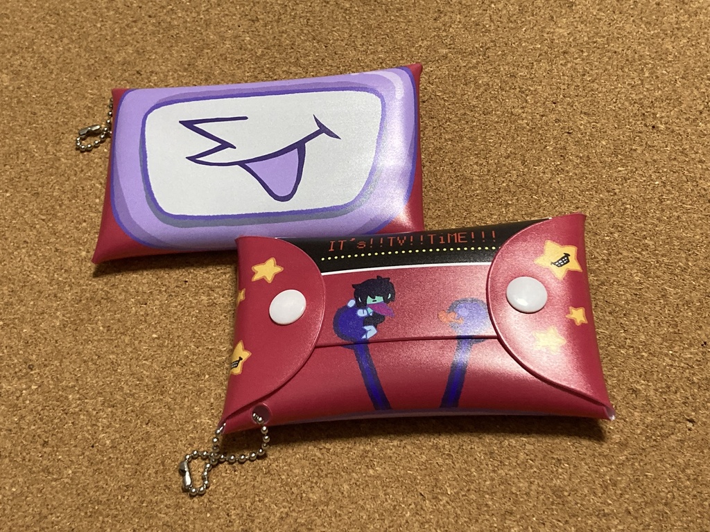 Two small plastic DELTARUNE-themed cases with printed faces and a ball chain on carpet.
