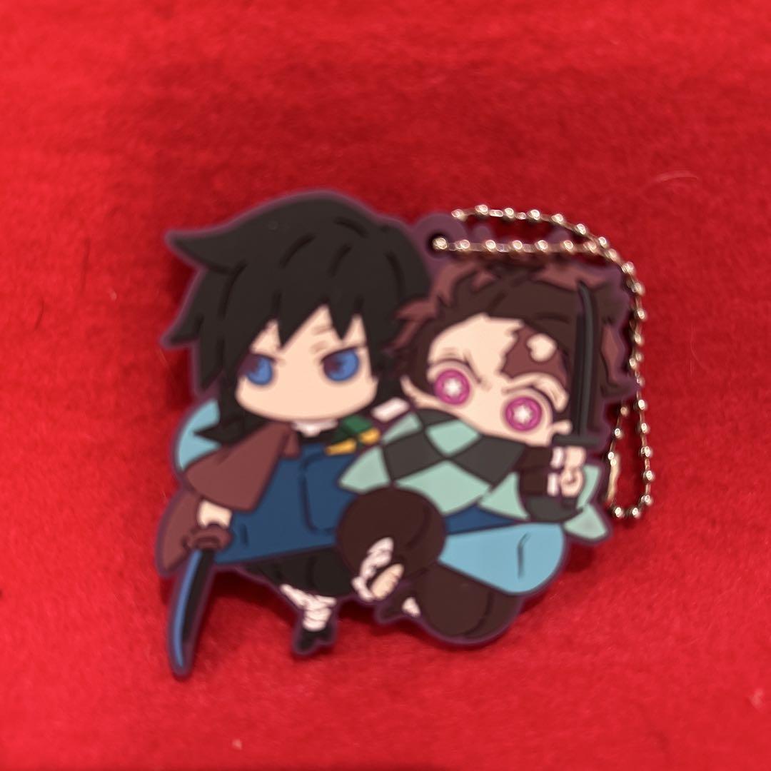 Front of a rubber keychain showing chibi Giyu and Tanjiro characters from Demon Slayer on a red background.