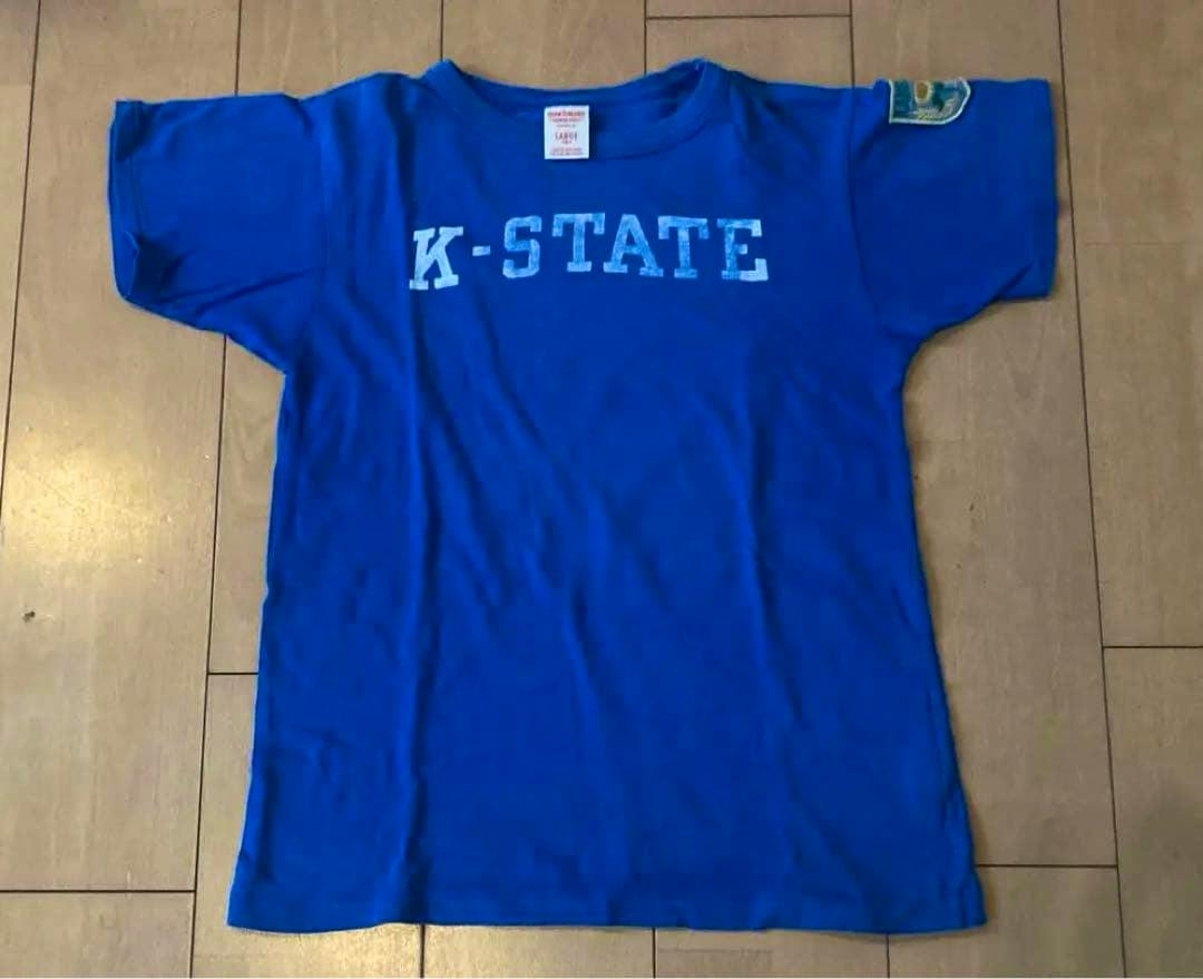 Blue children's cotton T-shirt laid flat on a wooden floor showing the front with “K-STATE” printed across the chest.