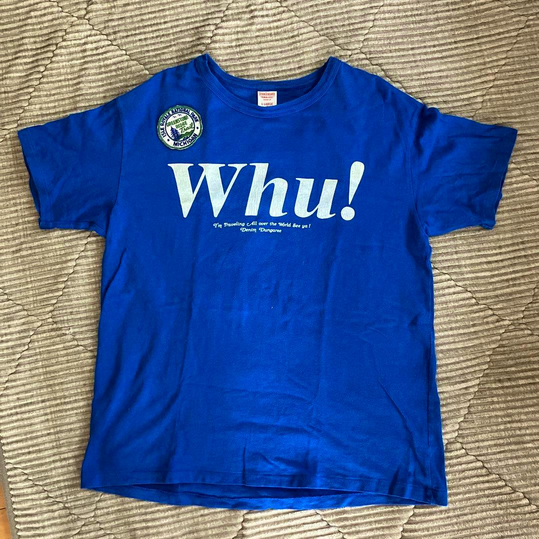 Blue short-sleeve T-shirt laid flat showing the front with a large white "Whu!" print and a small round patch on the left chest.