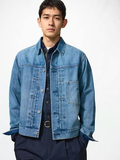 Male model wearing a light blue denim trucker jacket over a dark shirt on a white background.