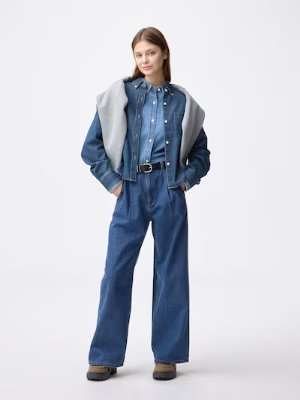 A full-length photo of a model wearing the denim tuck trousers with a denim shirt and jacket.