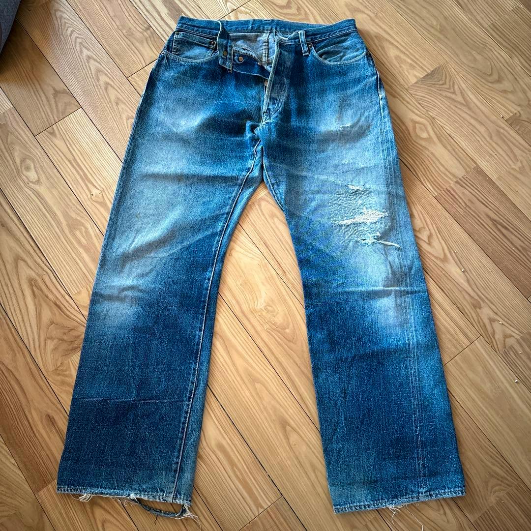 Front view of men's denim jeans laid flat on a wooden floor showing faded wear and a tear on the right thigh.
