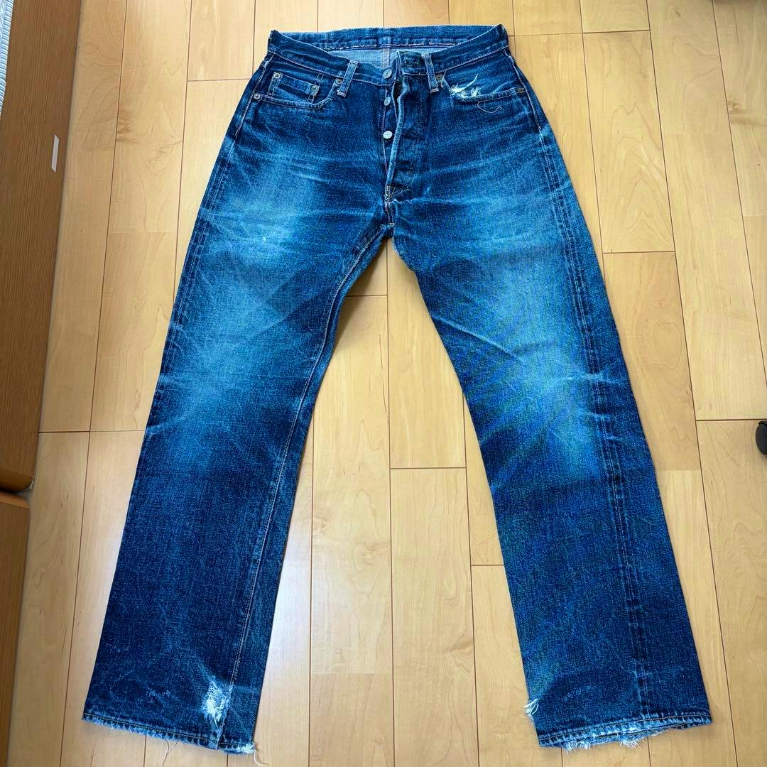 Front view of men's denim jeans laid flat on a wooden floor, showing fade patterns and distressed hems.