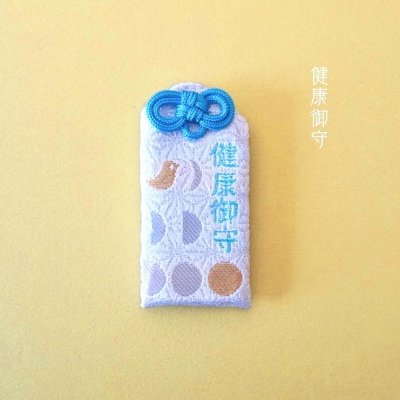 A pale-blue embroidered cloth health amulet (omamori) with a blue decorative knot photographed on a light background.