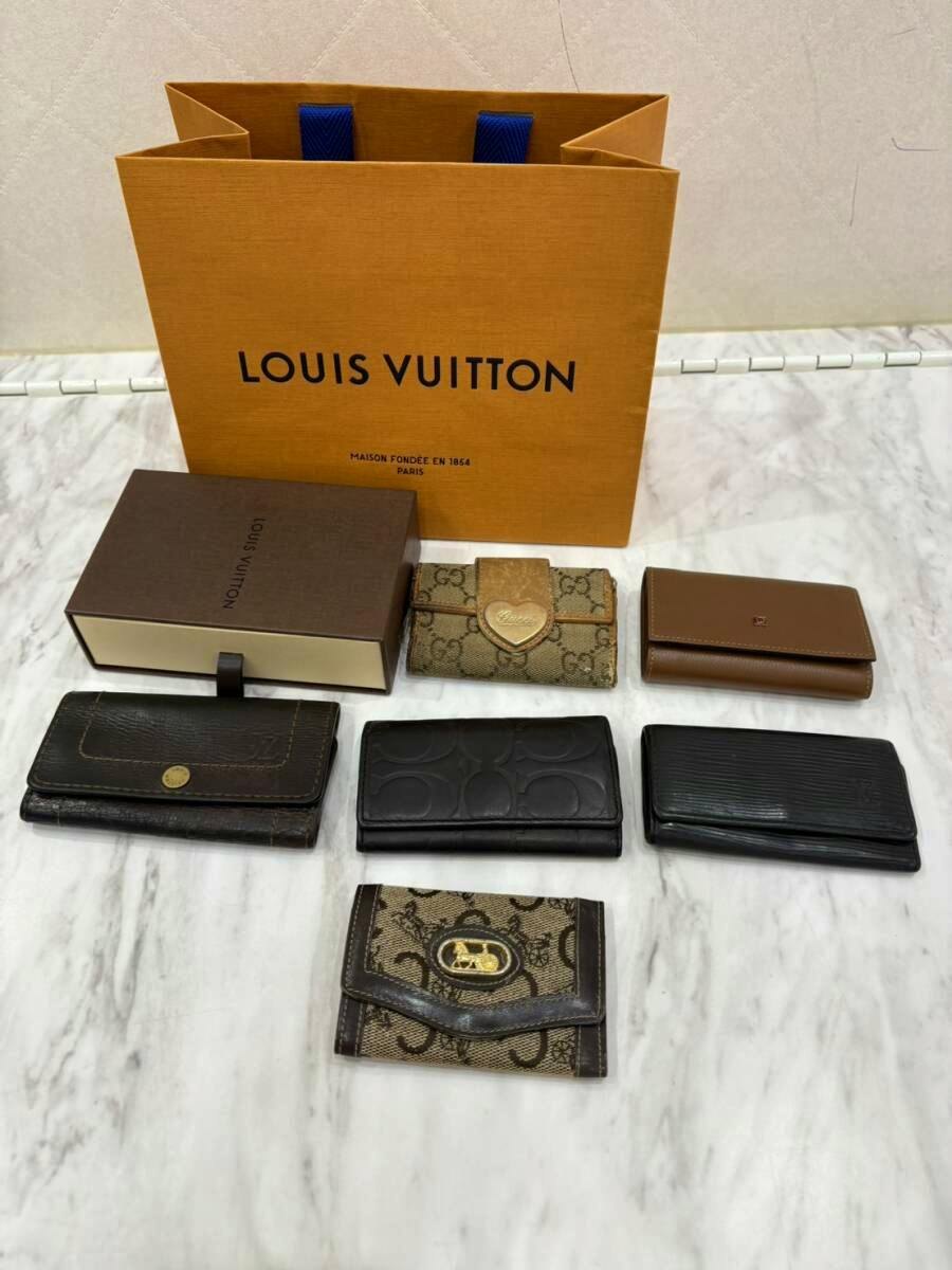 Six branded key cases arranged on a marble surface with a Louis Vuitton paper bag and a Louis Vuitton box behind them.