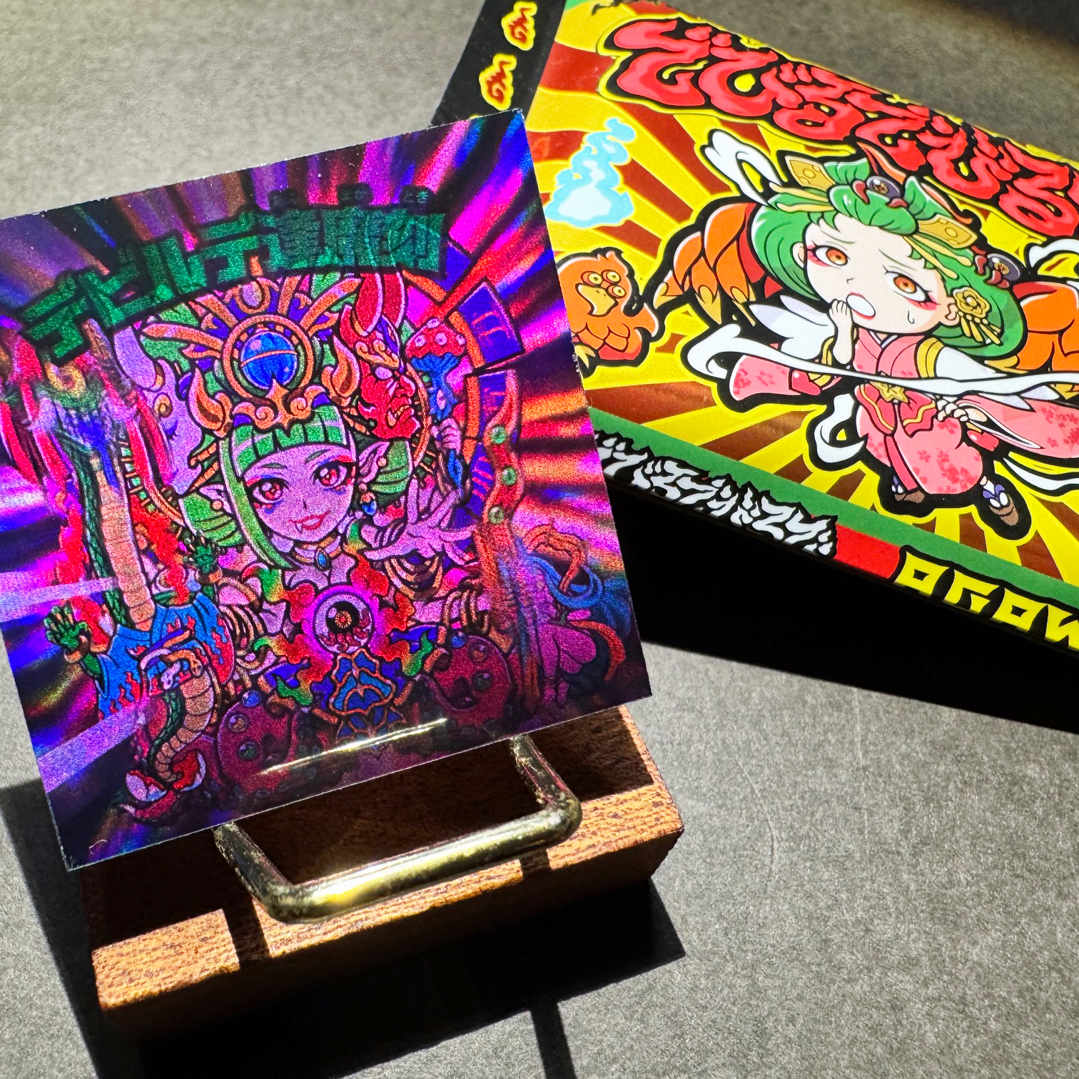 Holographic square sticker label with a colorful character illustration displayed on a small wooden stand next to its box.