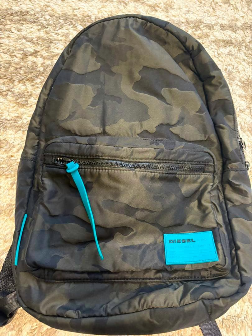 Front view of a camouflage-pattern DIESEL backpack showing a front zip pocket and a turquoise logo patch.