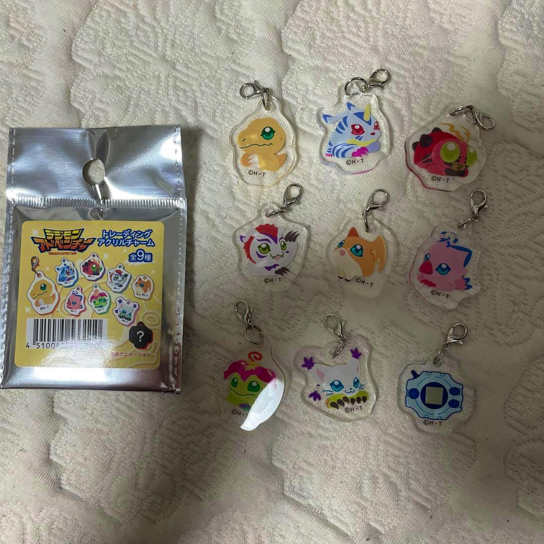 Several Digimon acrylic keychain charms with lobster clasps arranged next to a sealed foil package on a textured cloth.