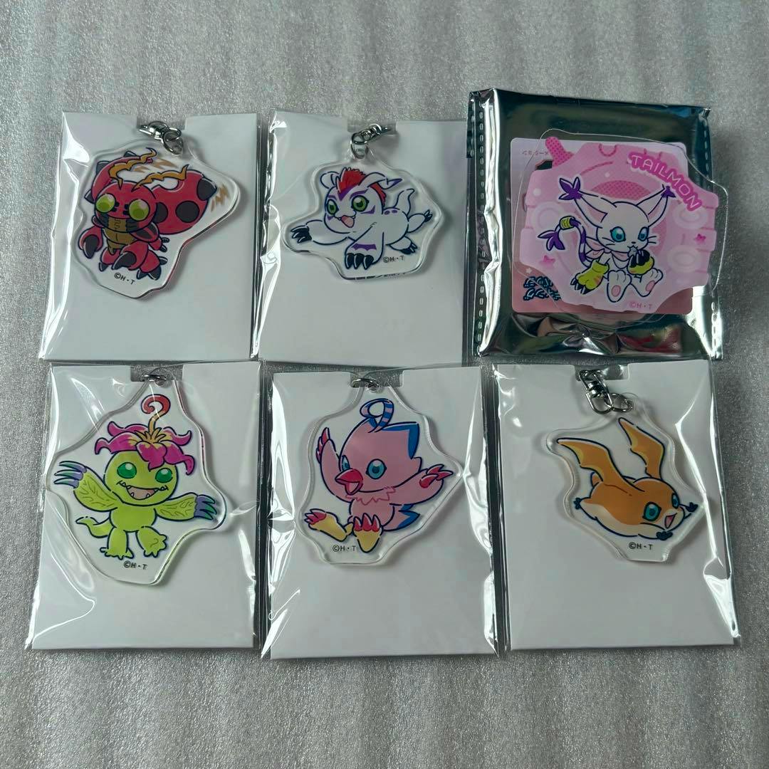 Several Digimon Adventure acrylic keychains and a packaged acrylic badge shown in individual plastic sleeves arranged on a fabric surface.