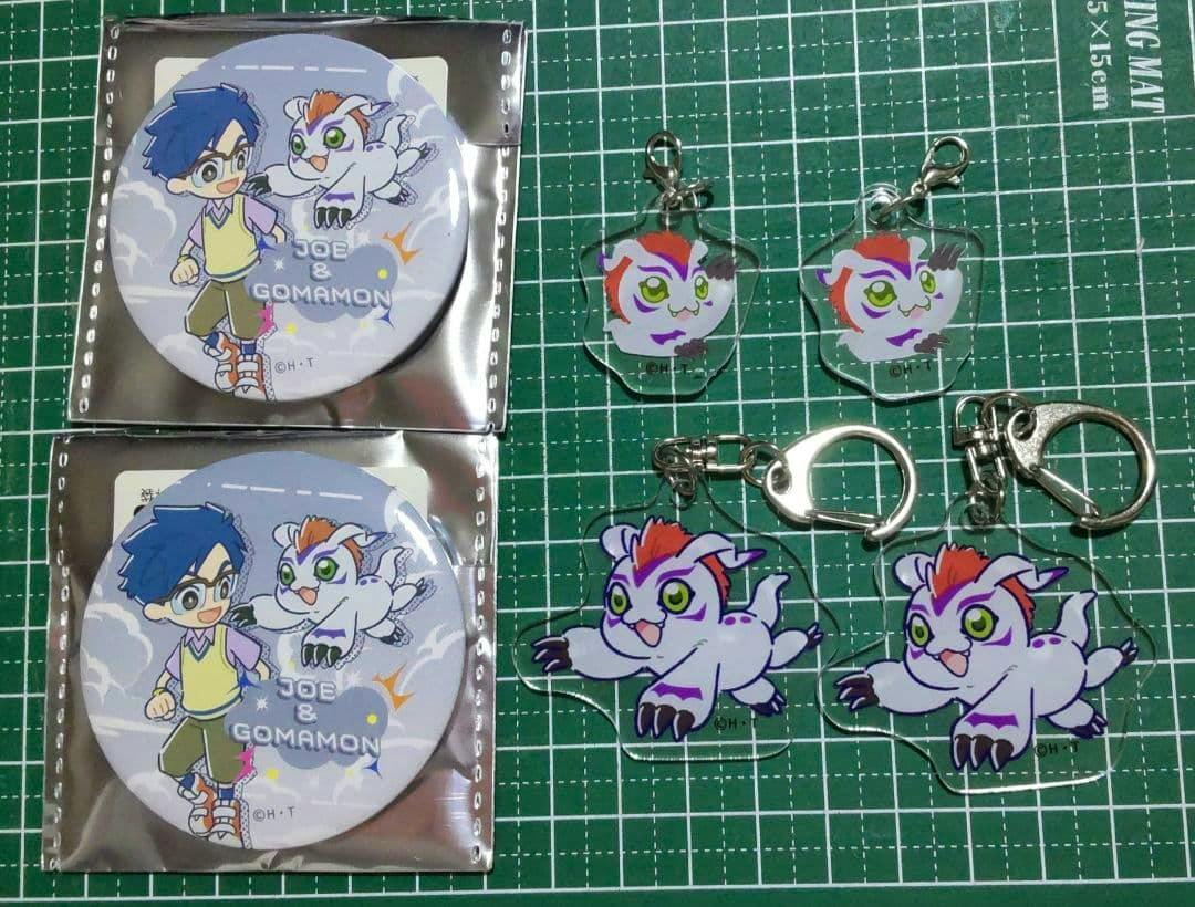 Two round can badges and four Gomamon acrylic keychains laid out on a green cutting mat.