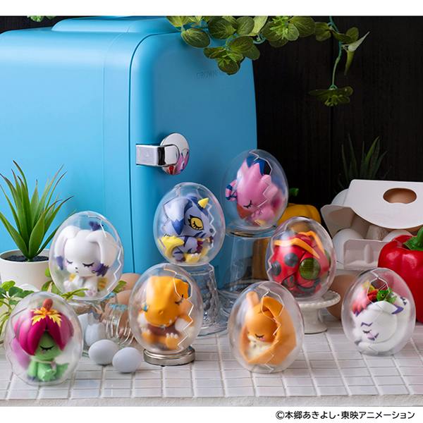 A set of Digimon MEGA EGG FIGURE plastic figures seated inside clear egg-shaped capsules arranged on a tabletop in front of a small blue mini-fridge.