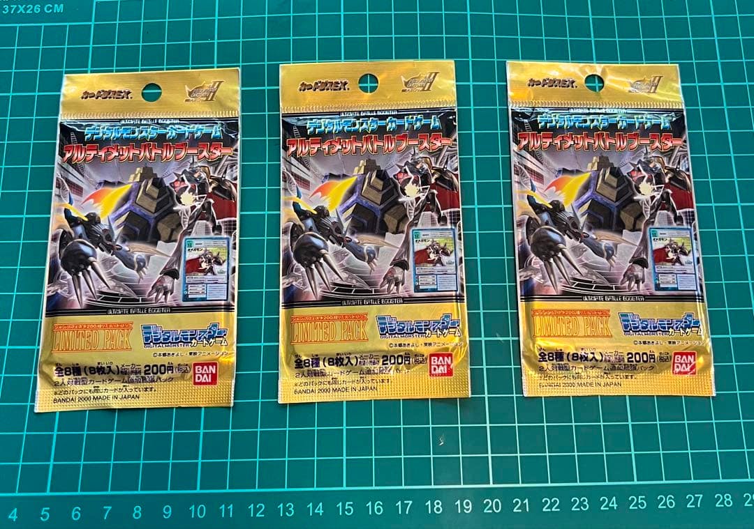 Three sealed Digimon Card Game "Ultimate Battle Booster" packs showing the front packaging laid on a green cutting mat.