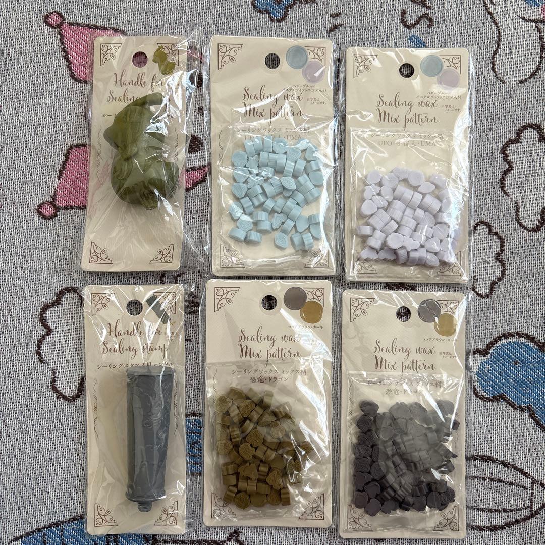 Photo of six packaged items for a sealing wax set: a dinosaur-shaped stamp handle, a second stamp handle, and four packs of sealing wax beads on a patterned fabric background.