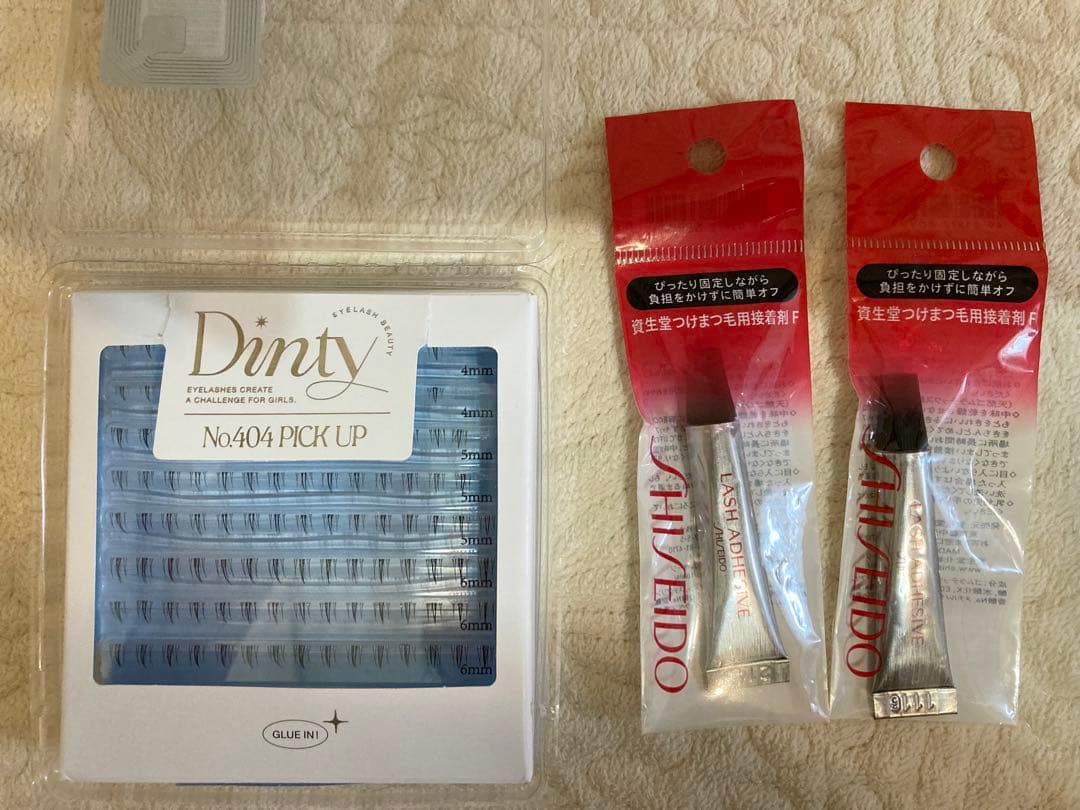 A tray of Dinty No.404 lower false eyelashes on the left and two packaged Shiseido eyelash adhesives on the right on a light textured surface.
