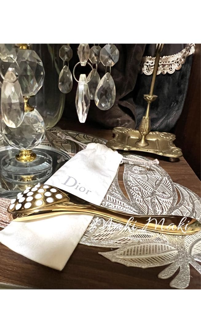 Gold Dior face roller with a multi-nubbed rolling head resting on a white Dior pouch on a wooden surface next to a crystal lamp.