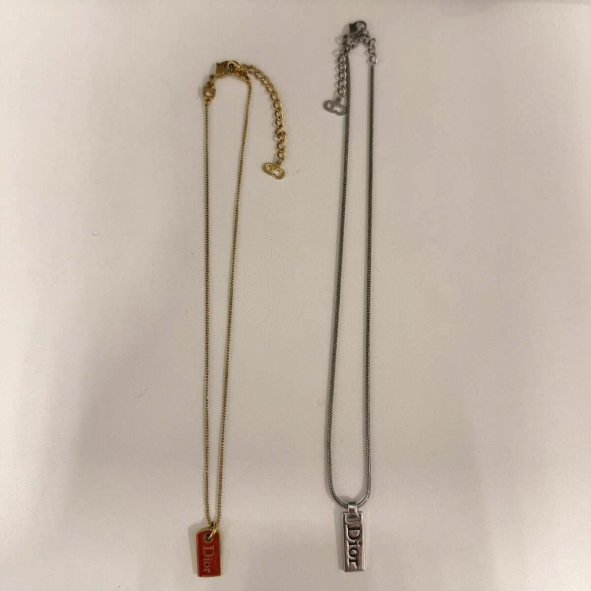 Two necklaces laid flat on a white background: a gold-tone chain with a small red rectangular Dior pendant and a silver-tone chain with a silver rectangular Dior pendant.