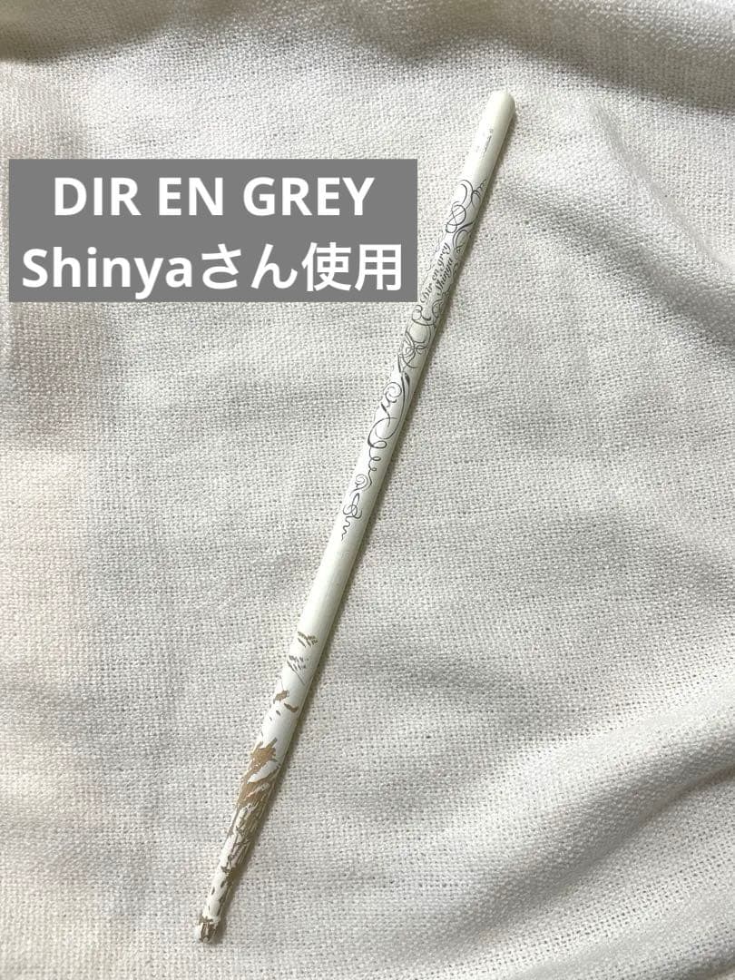 A single white drum stick with decorative black script and a chipped tip lying on white fabric, with overlaid Japanese and English text.