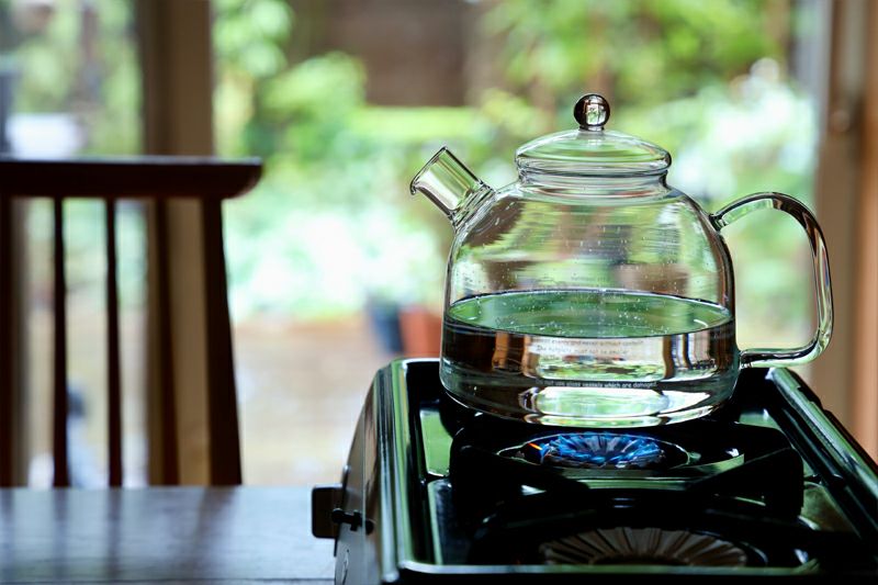 A clear glass tea kettle on a gas stove partially filled with water with a blue flame beneath.