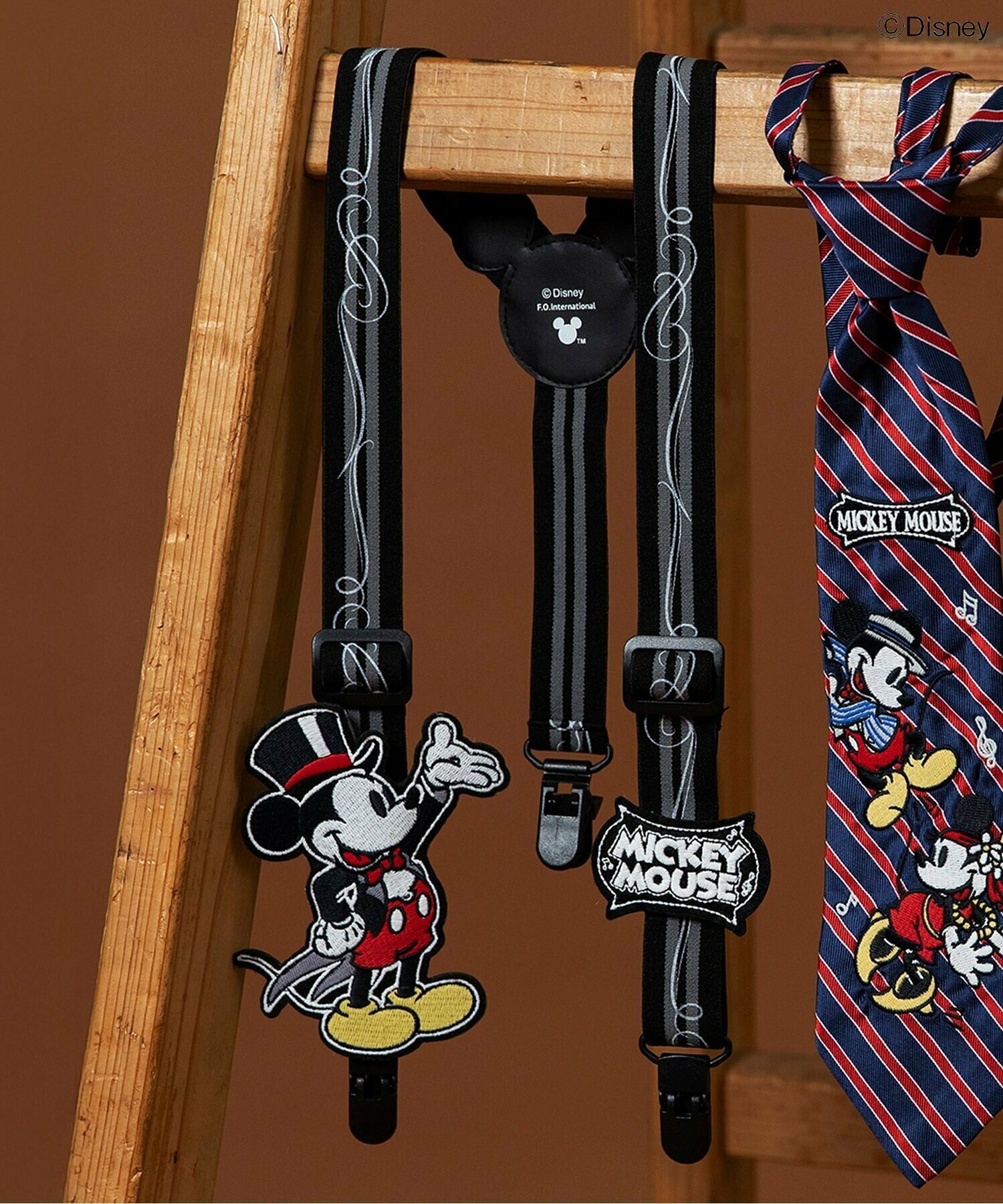 Black striped suspenders with an embroidered Mickey Mouse character patch and a 'MICKEY MOUSE' logo hanging on a wooden ladder.