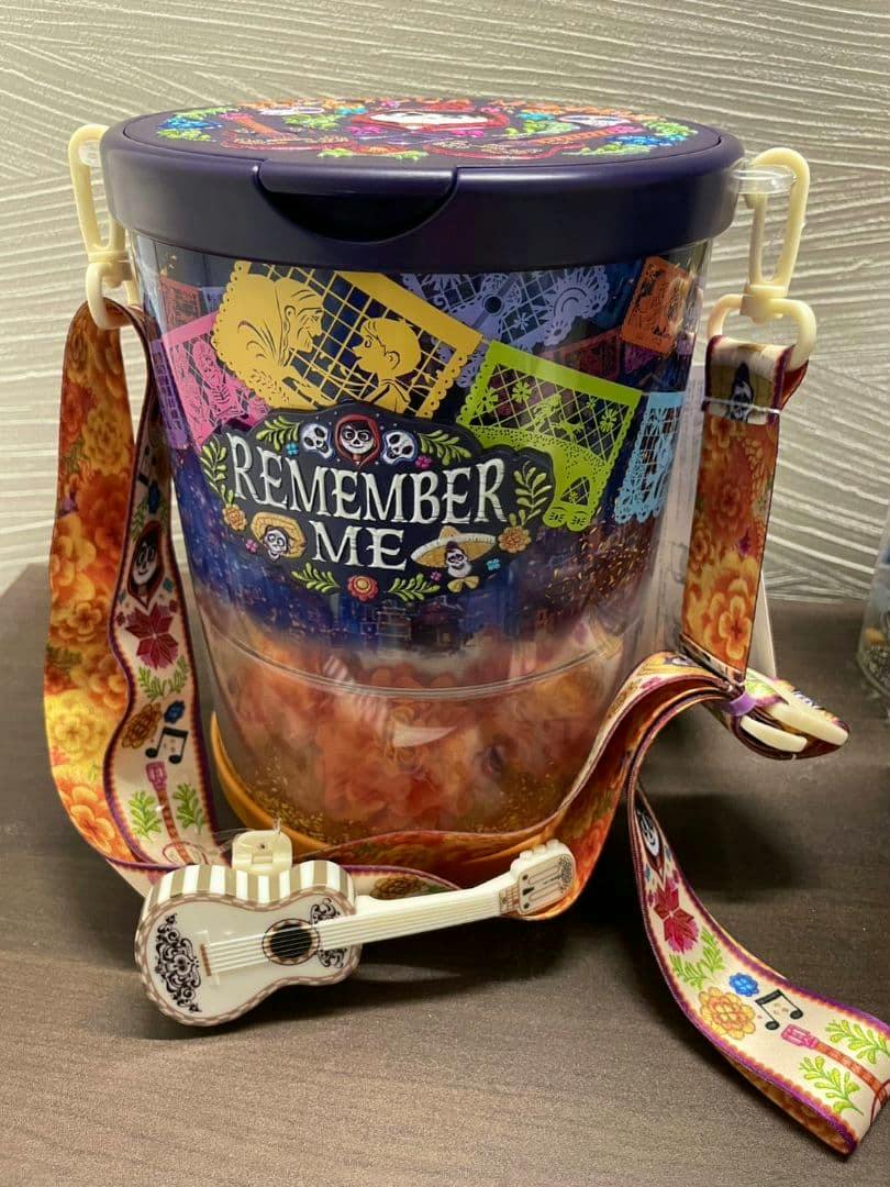 Plastic Disney "Remember Me" popcorn bucket with a purple lid, clear decorated body, patterned shoulder strap, and a small guitar charm.