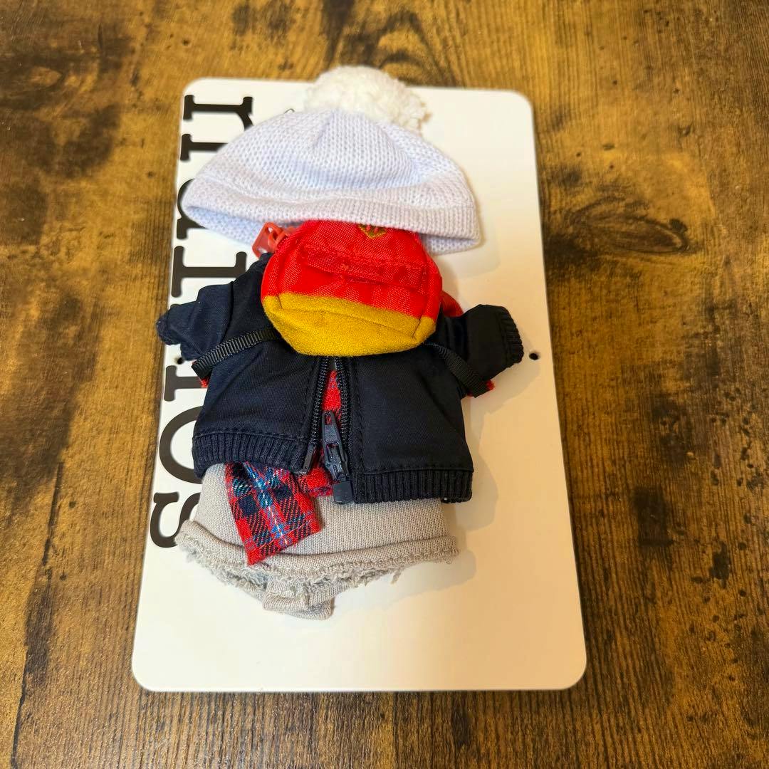 Top-down photo of a set of doll clothes including a white knit hat, a navy zip jacket, a small red and yellow backpack, a red plaid scarf, and gray bottoms arranged on a white card.