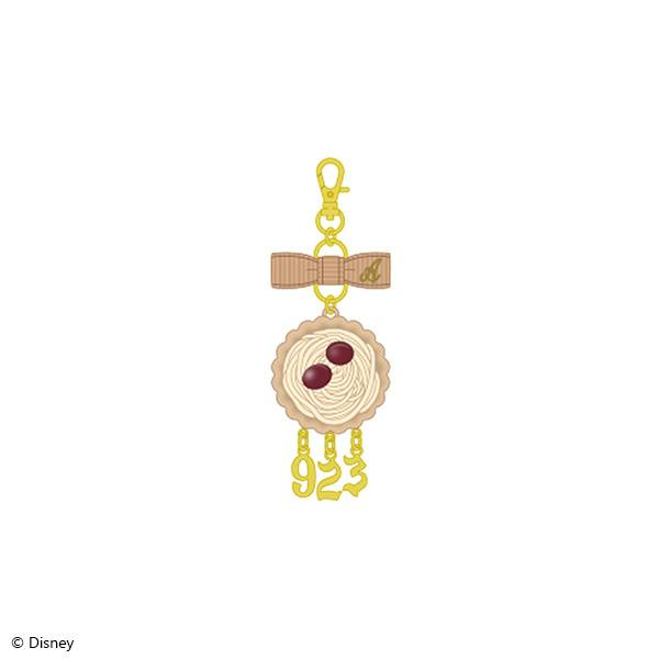 Front view of the Ace Trappola La・Bonbonniere version brooch charm keychain showing a bow, a round cream-colored pastry charm with two red beans, and three dangling gold number charms.