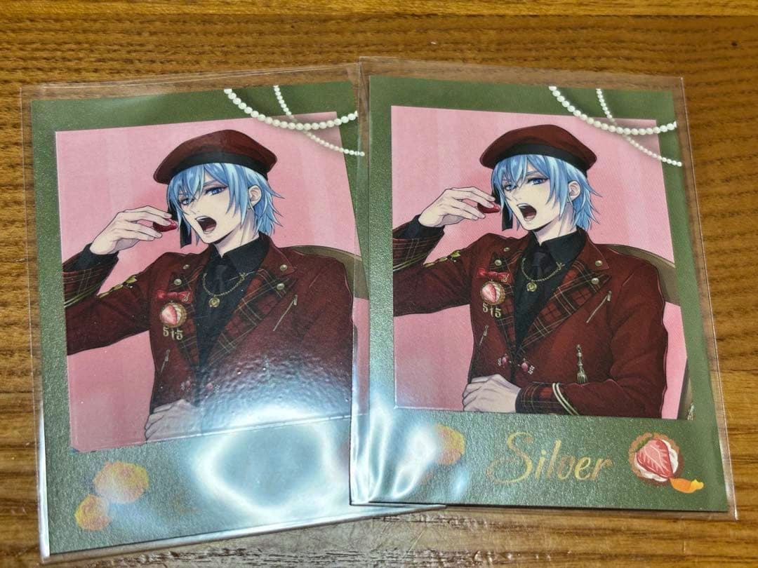 Two photocards in plastic sleeves showing a blue-haired illustrated character in a red outfit with "Silver" printed on the card, placed on a wooden surface.
