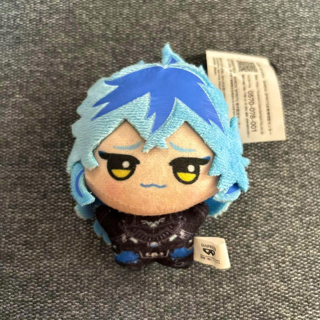 Front view of a small blue-haired chibi plush toy (イデア) with embroidered facial features and attached tags on a gray fabric surface.