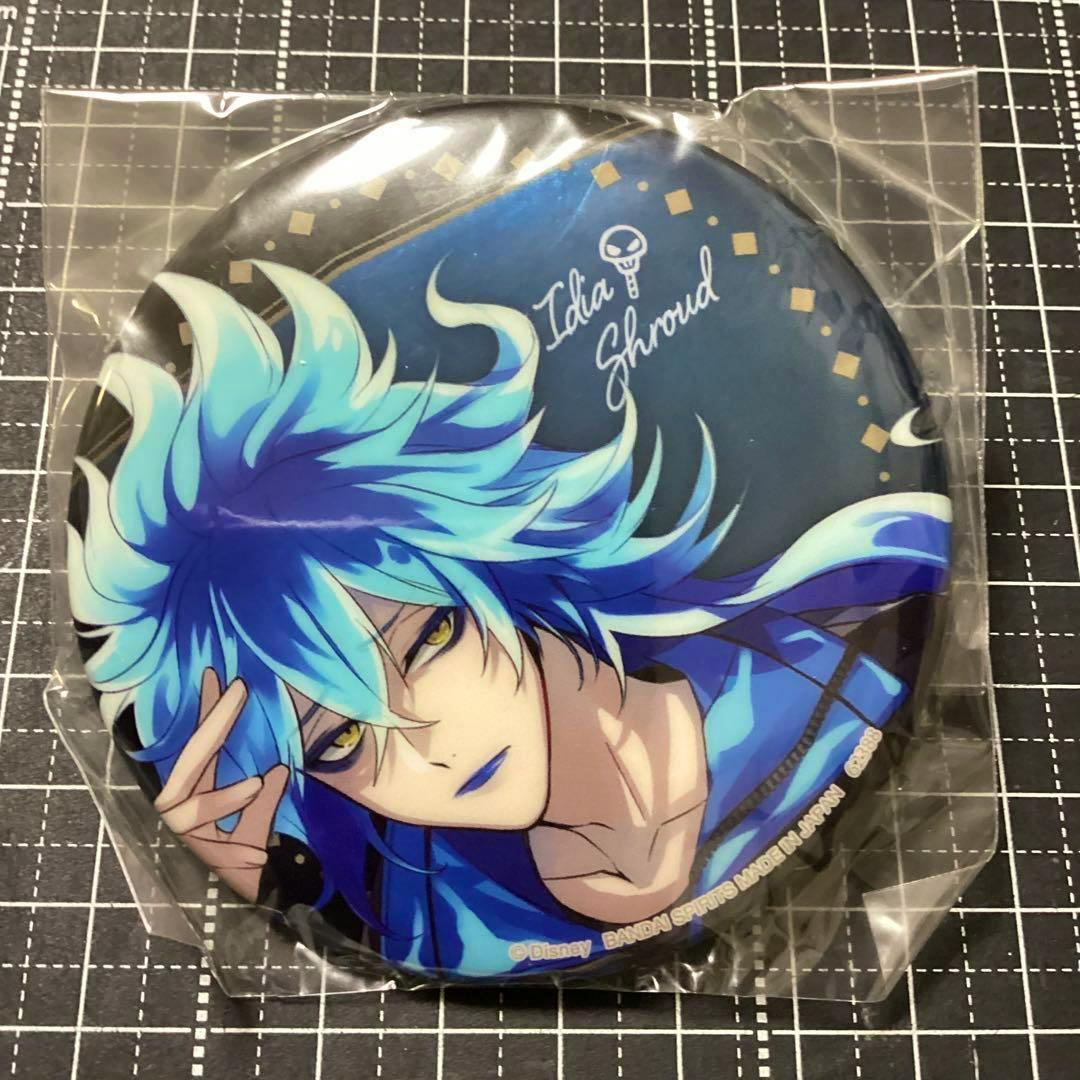 Photograph of a round Twisted Wonderland (一番くじ) can badge showing the character イデア/Idia with blue hair, in its plastic wrapper on a gridded background.