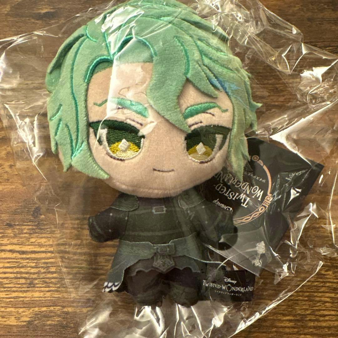 A Sebek Twisted Wonderland plush in its clear plastic packaging, showing green hair and a dark outfit on a wooden surface.