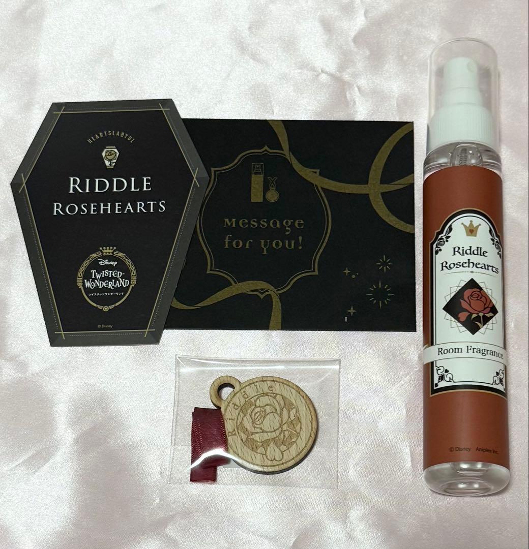 A plastic room fragrance spray bottle labeled “Riddle Rosehearts” pictured alongside packaging cards and a small wooden charm.