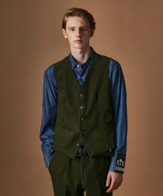 Male model wearing a green buttoned vest over a blue long-sleeve shirt.