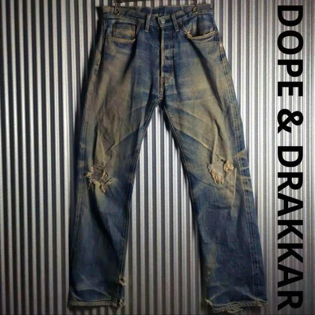 Front view of men's distressed denim jeans with knee rips hung on a corrugated metal background.
