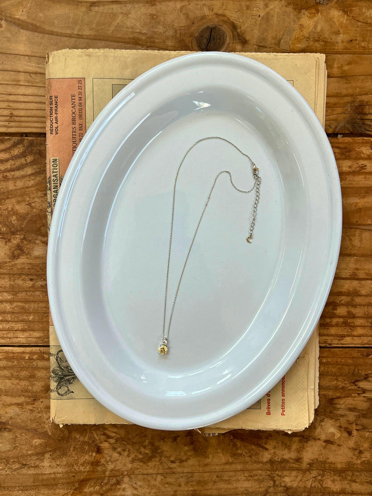 Top-down photo of the Double Stone Lemon Necklace laid on a white oval dish showing the chain and small yellow double-stone pendant.