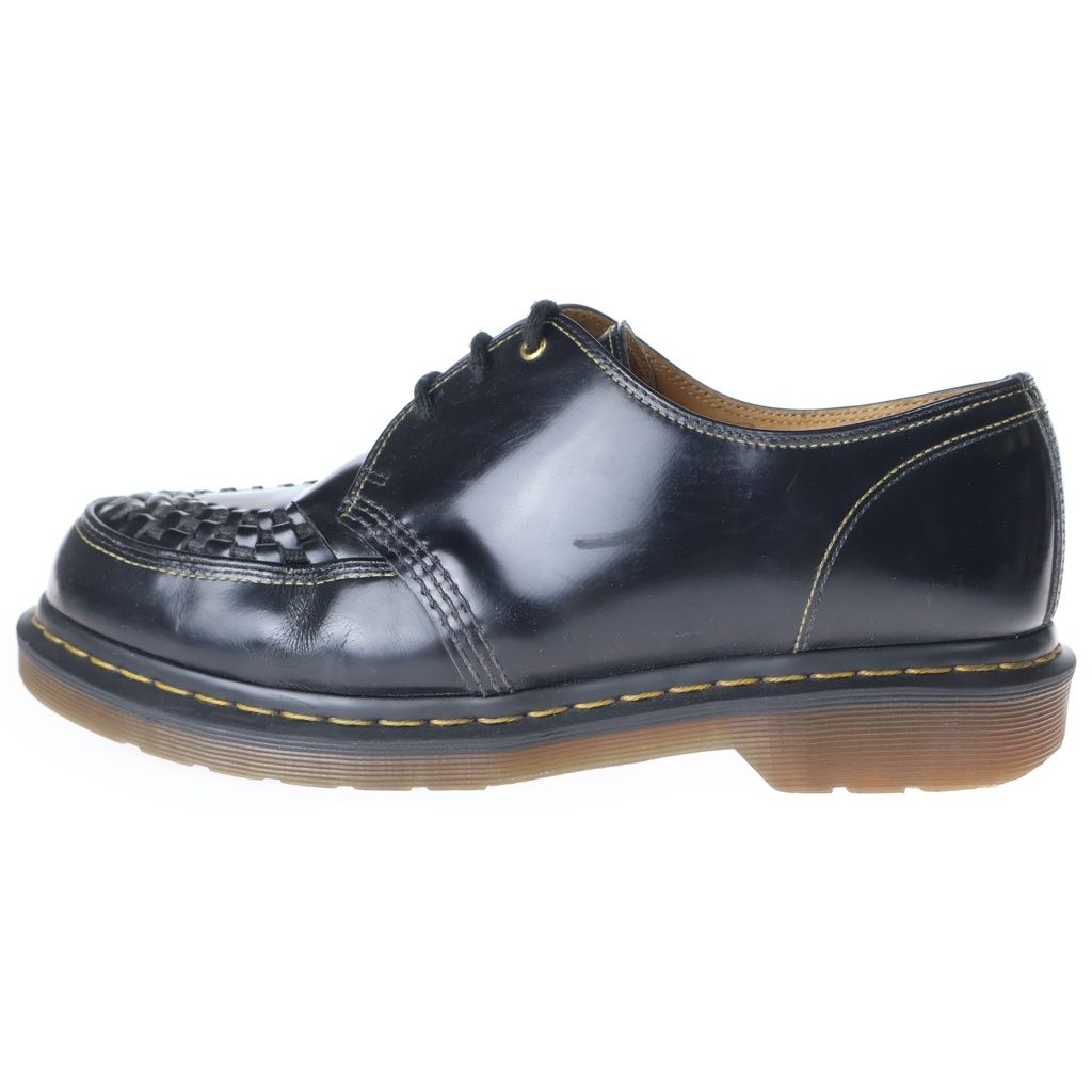 Side view of a black leather Dr. Martens three-hole shoe showing woven toe, yellow welt stitching, and translucent sole.