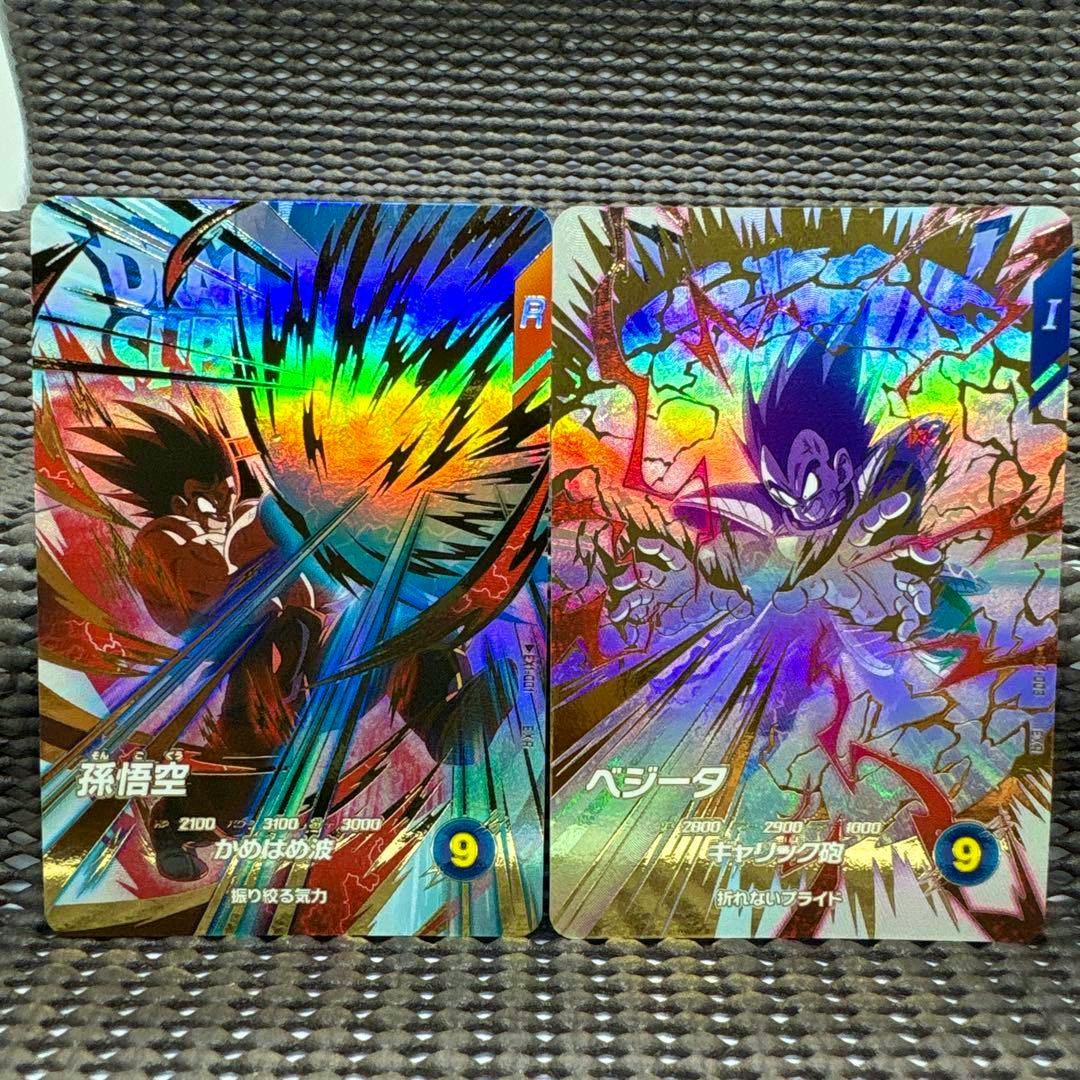 Two holographic trading cards showing the front artwork of Son Goku and Vegeta side by side on a textured surface.