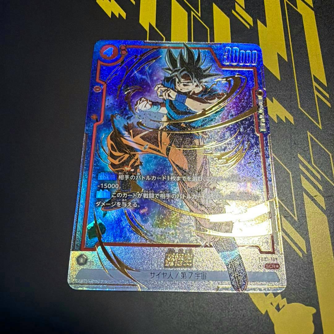 Photograph of the trading card featuring 孫悟空 (Goku) with holographic foil, placed on a black mat with gold geometric designs.