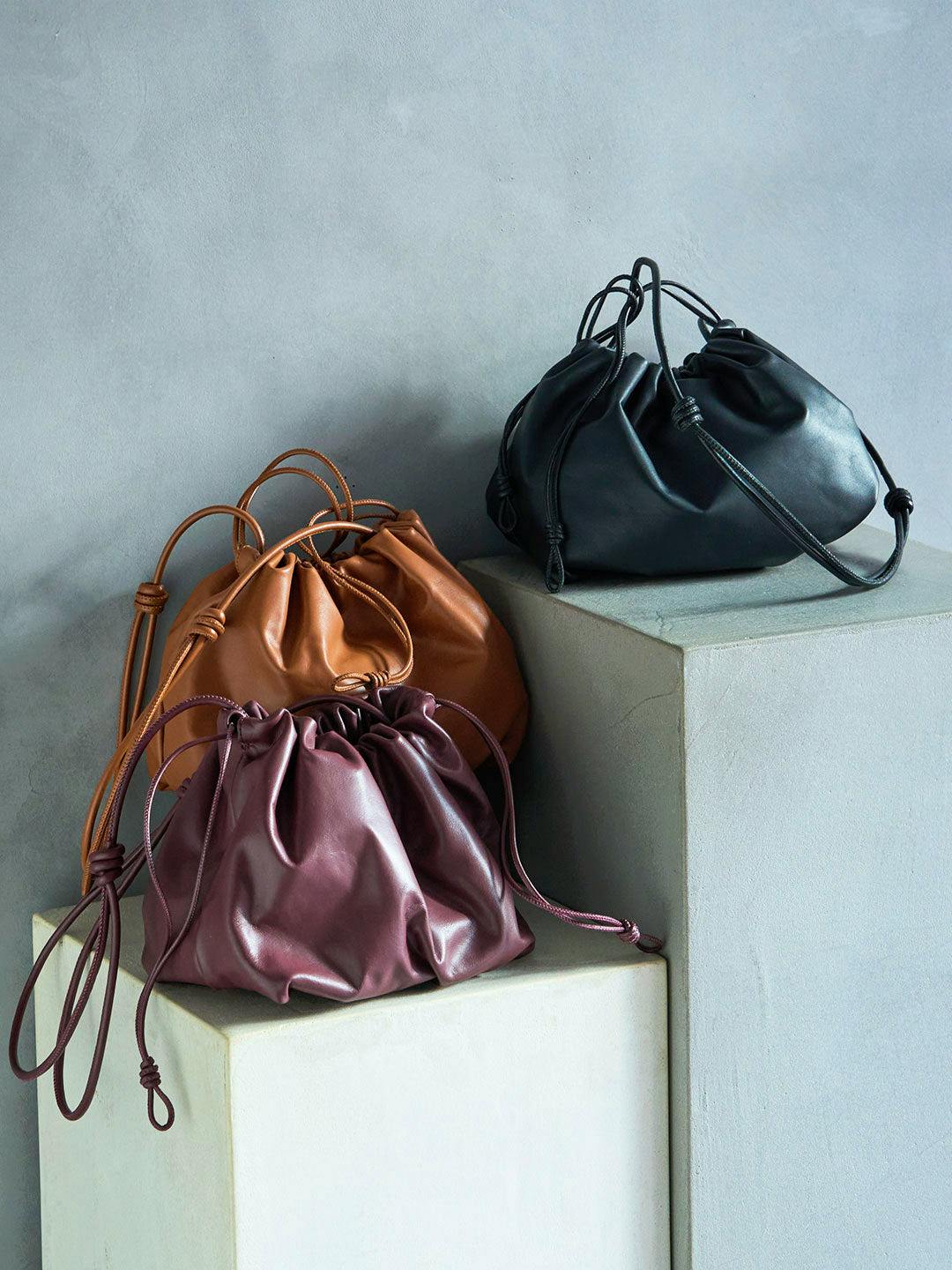 Three drawstring leather shoulder bags in black, brown, and burgundy displayed on gray pedestals.