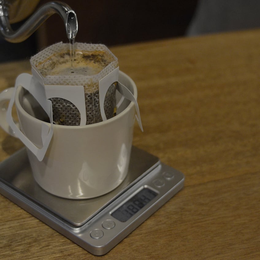 A drip bag coffee filter mounted on a white mug with hot water being poured into it on a digital scale.