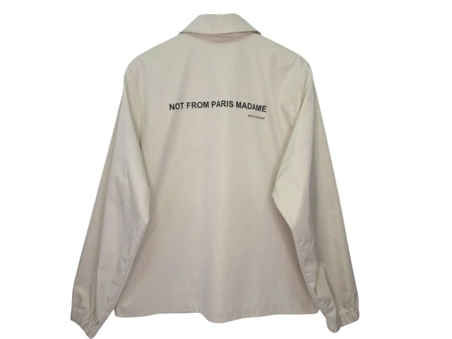 Beige long-sleeve jacket photographed from the back showing the printed text "NOT FROM PARIS MADAME" on a plain background.