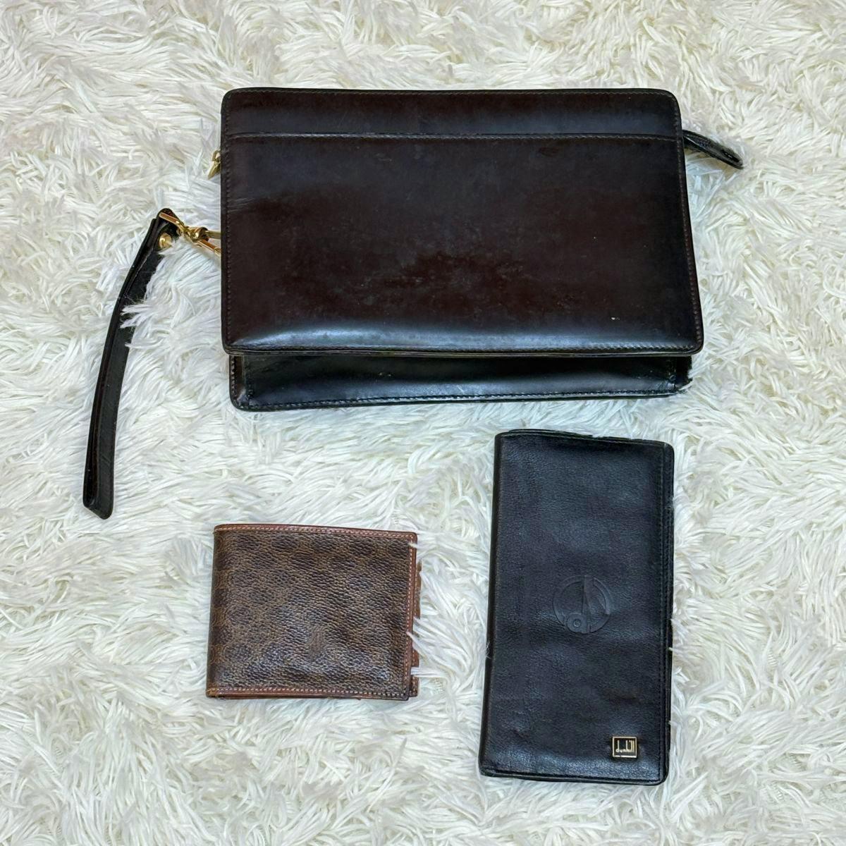 A dark brown leather clutch bag with a wrist strap is shown alongside two leather wallets on a white shag rug.